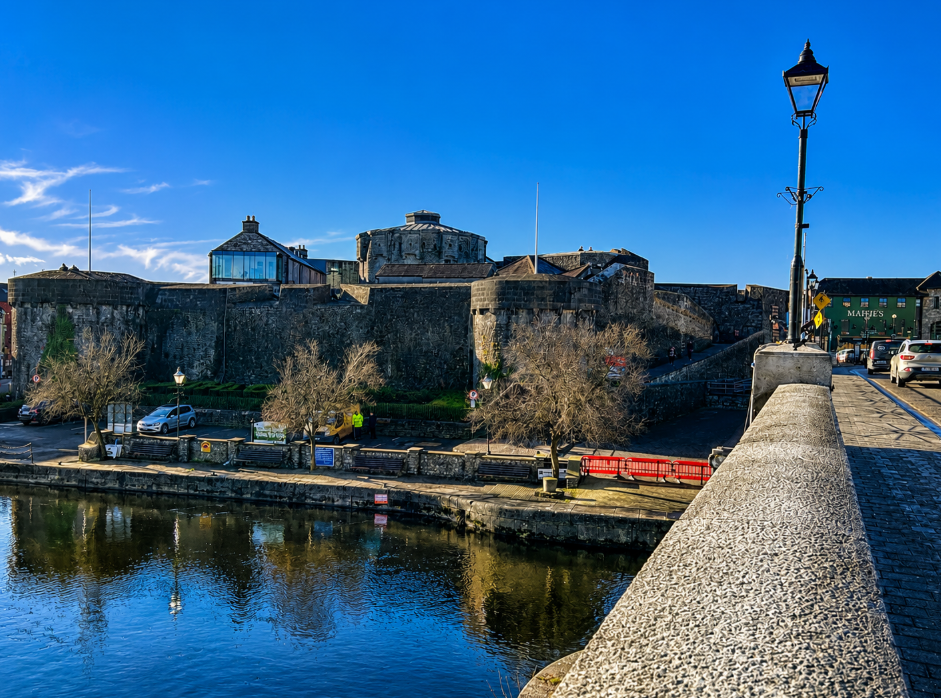 Athlone Castle viewed from across the River Shannon in Athlone, Ireland, with the historic stone fortress reflected in the water beneath a bright blue sky.