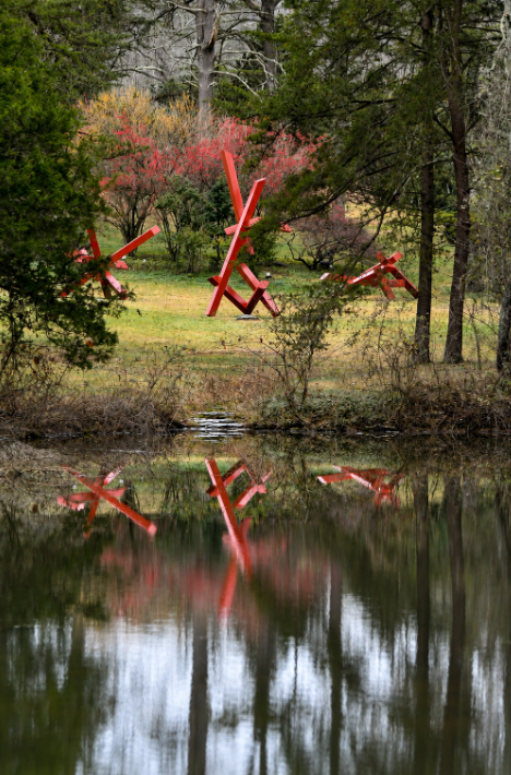 Bright red abstract sculptures reflected in a pond, framed by trees and spring foliage at Bernheim Forest and Arboretum in Kentucky.