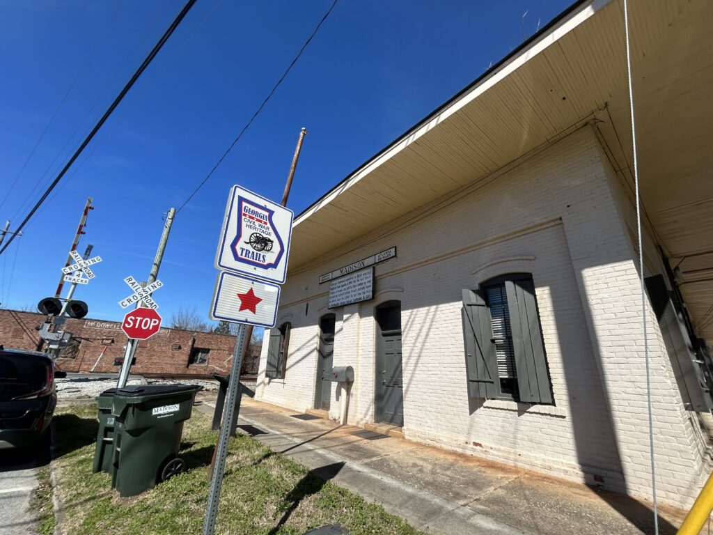 Exterior view of the historic Madison Station building in Madison, Georgia, part of the Georgia Civil War Heritage Trails. The photo includes the cream-colored brick depot with black shutters, a Georgia Civil War Trails marker with a red star, and nearby railroad crossing signs and tracks under a clear blue sky.