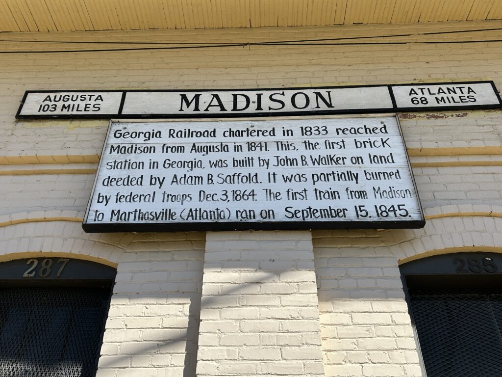 Historic sign on the Madison train depot in Madison, Georgia, displaying railroad history. The sign notes that the Georgia Railroad reached Madison from Augusta in 1841 and that the first brick station in Georgia was built here by John B. Walker. It also mentions that the depot was partially burned by federal troops on December 3, 1864, and that the first train from Madison to Marthasville (now Atlanta) ran on September 15, 1845. Directional markers indicate Augusta is 103 miles away and Atlanta is 68 miles away.