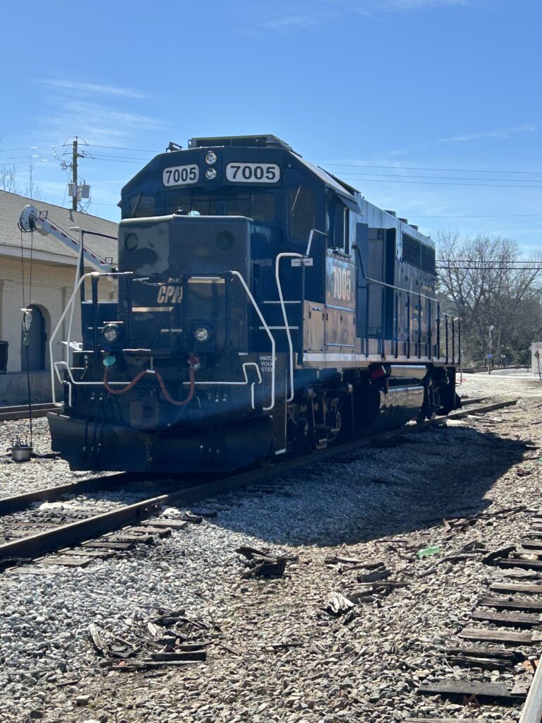 Blue locomotive engine number 7005 from the Central of Georgia Railway (CGR) sitting on the tracks near the historic Madison Station in Madison, Georgia, under a clear blue sky. The engine is positioned facing the camera with gravel-covered tracks and railroad remnants visible in the foreground.