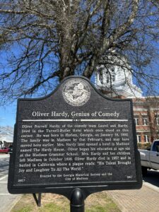 Historical marker titled "Oliver Hardy, Genius of Comedy" located in Madison, Georgia. The plaque details Oliver Hardy's early life, noting that his family moved from Harlem, Georgia, to Madison in early 1892. They lived at the Turnell-Butler Hotel, and his mother later ran the Hardy House boarding house. The marker also states Hardy began school at Madison Grammar School before the family left in 1898. Erected by the Georgia Historical Society and the City of Madison, the plaque honors Hardy’s legacy with the quote: “His talent brought joy and laughter to all the world.”
