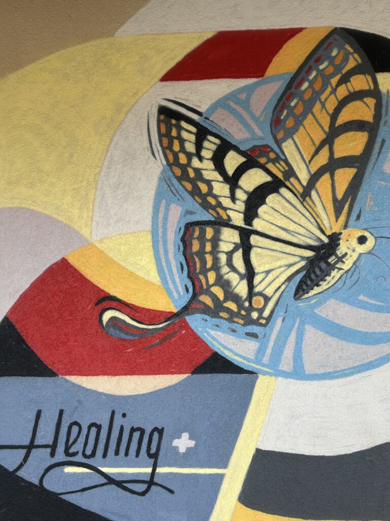 Colorful mural featuring a yellow and black butterfly in flight against abstract swirls of red, yellow, blue, and white. The word 'Healing' is written in elegant script near the bottom with a white medical cross symbol.