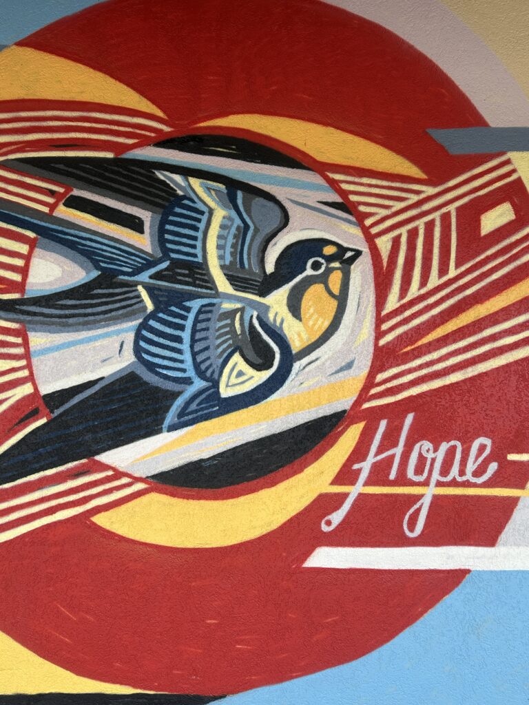 Vibrant mural of a bird in mid-flight with layered wings in blue, yellow, and black tones. The background includes bold red and yellow arcs, with the word 'Hope' written in white script to the right.