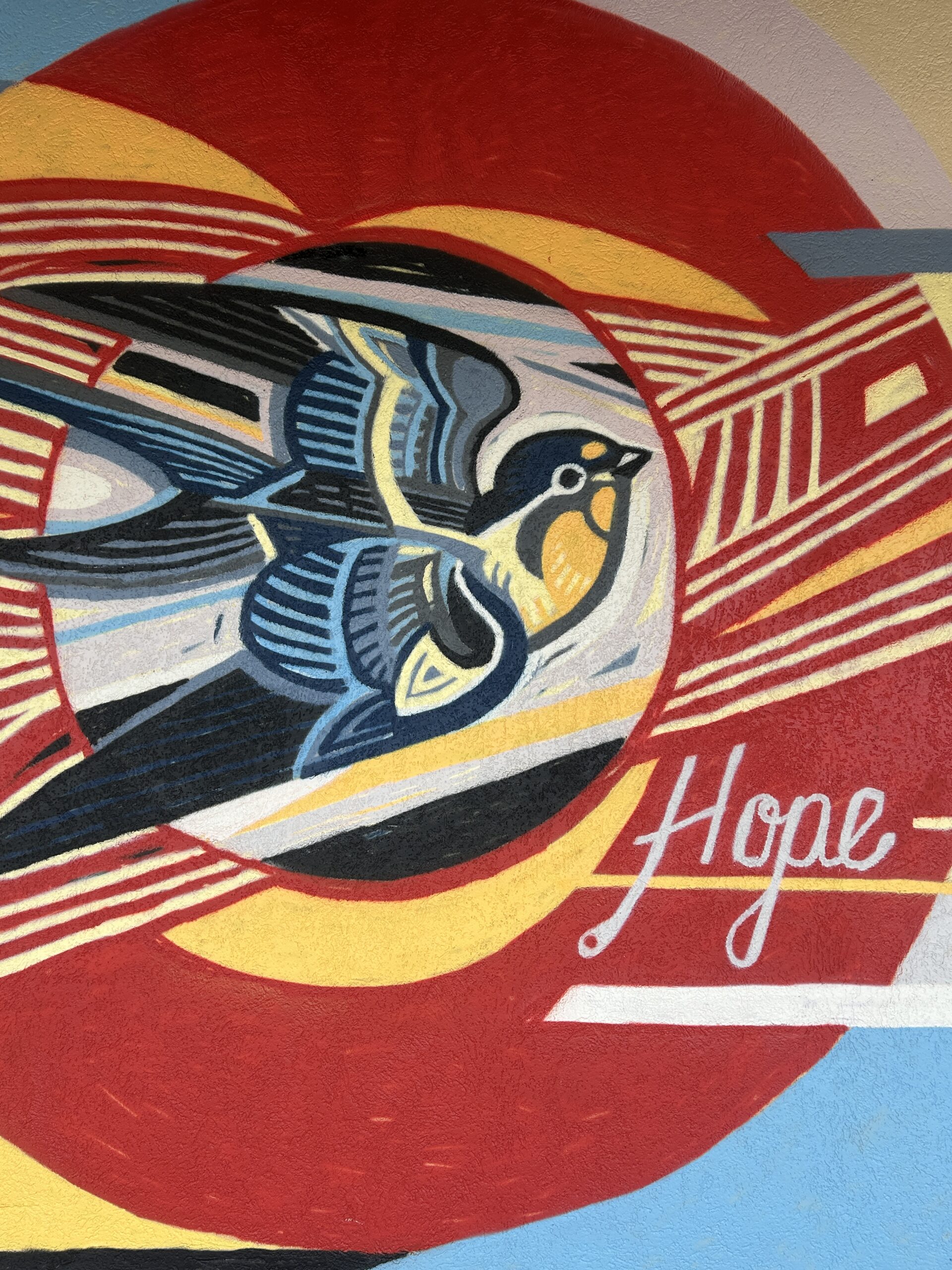 Vibrant mural of a bird in mid-flight with layered wings in blue, yellow, and black tones. The background includes bold red and yellow arcs, with the word 'Hope' written in white script to the right.