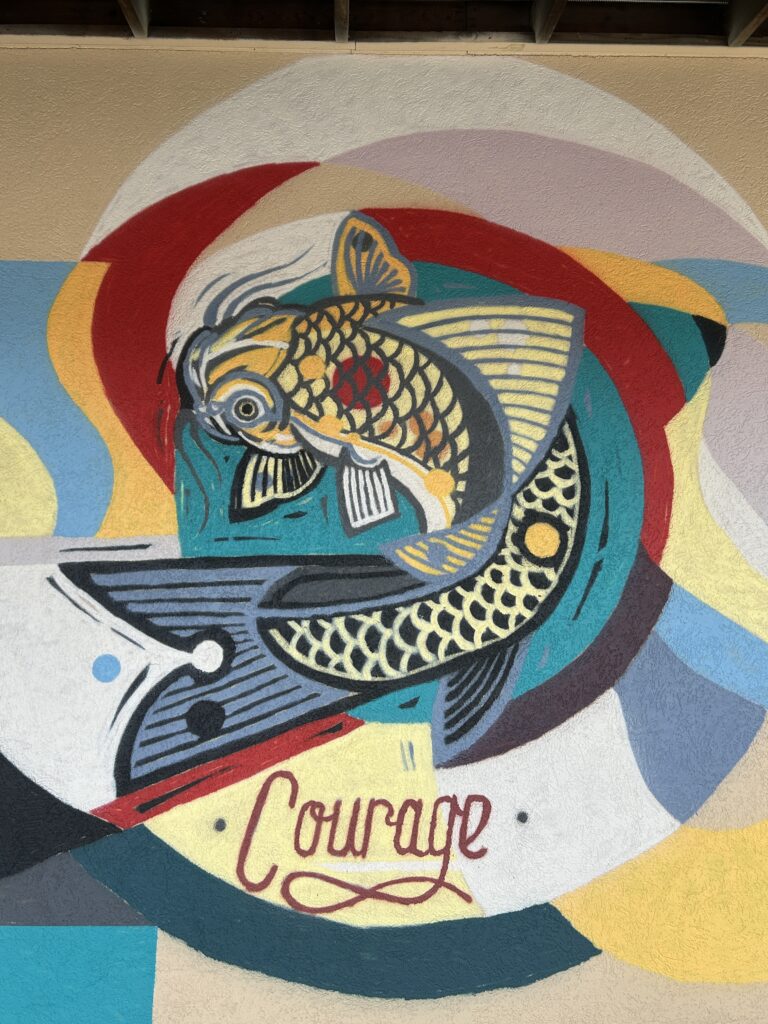 Street art mural featuring a golden koi fish swimming upward through waves of red, teal, and yellow. The word 'Courage' is written in red script below the fish, symbolizing strength and perseverance.
