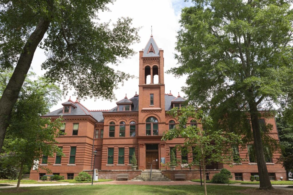 Front view of the Madison-Morgan Cultural Center in Madison, Georgia, a historic red-brick building with Romanesque architectural features, arched windows, and a central bell tower. Surrounded by large trees and green lawns, the structure was formerly Madison Grammar School, where Oliver Hardy began his education.