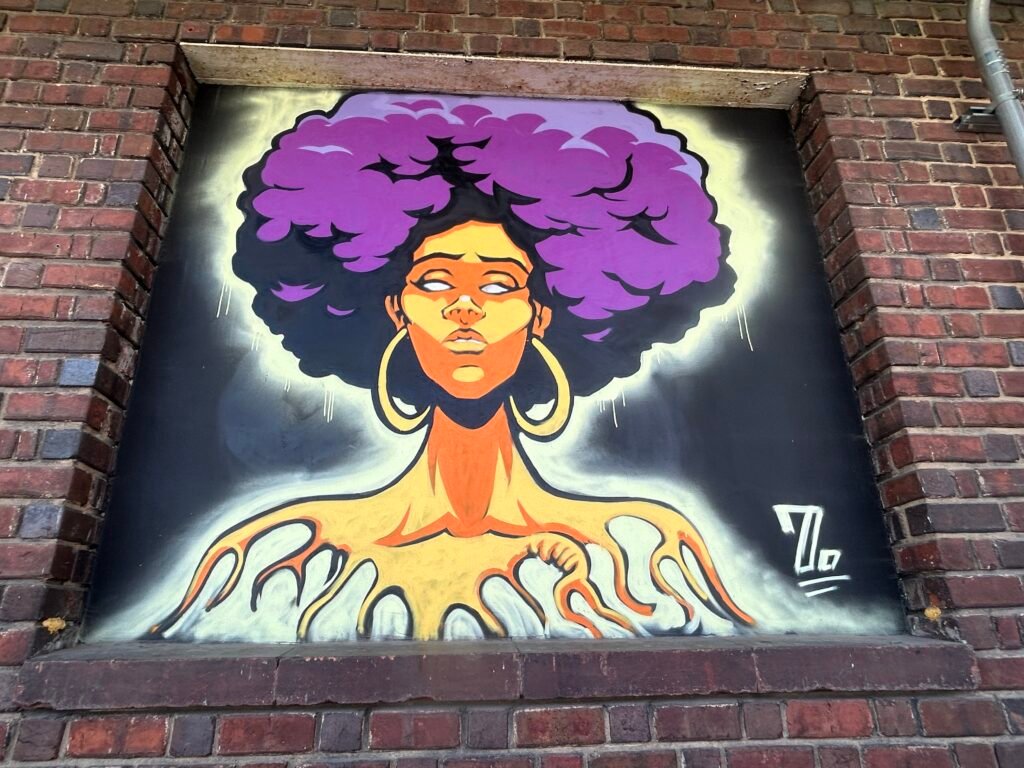 Portrait mural of a woman with voluminous purple hair, gold hoop earrings and hands reaching across her shoulders.