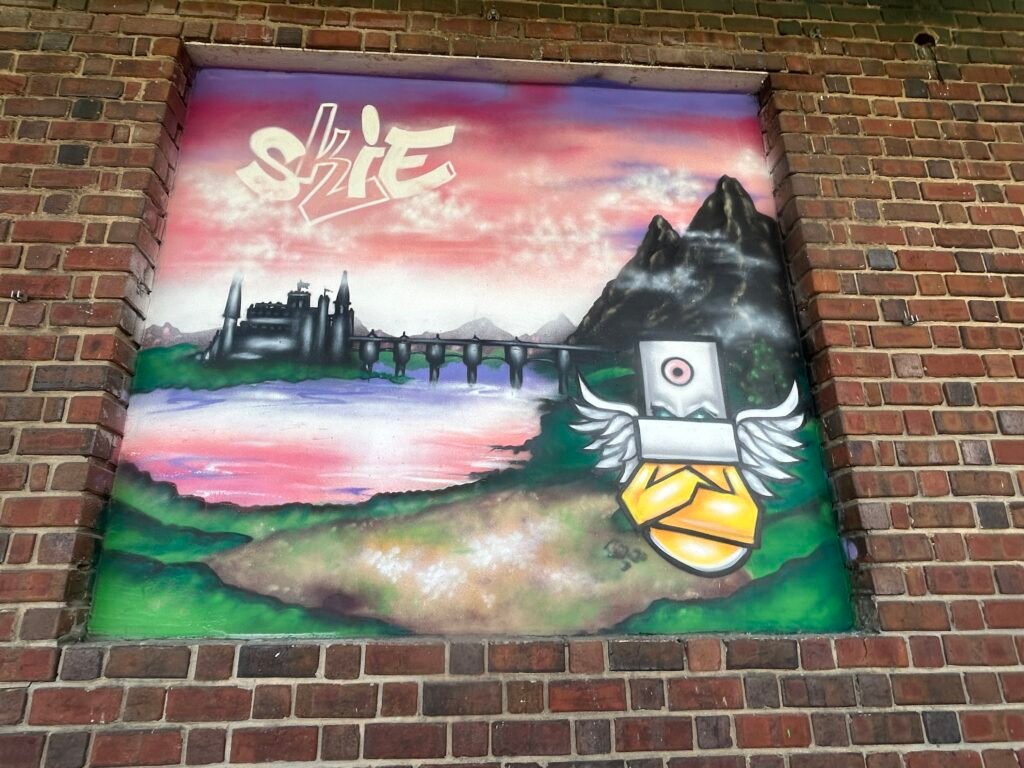 Winged, box-headed robot standing before a castle, bridge and mountains beneath a pink sky.