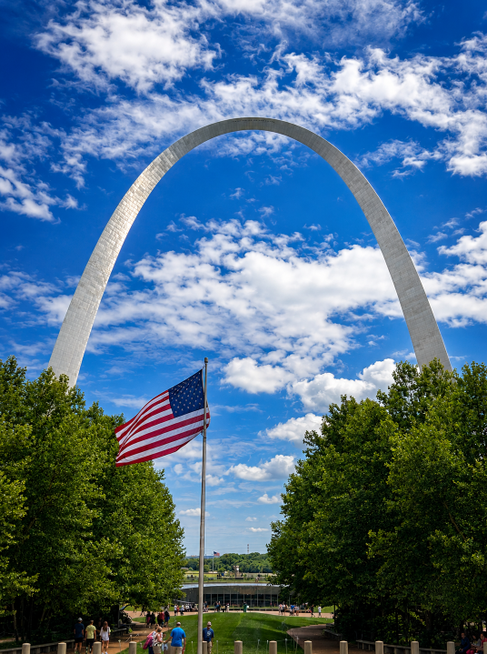 American flag framed beneath the Gateway Arch above the Reflection Pond on a bright, partly cloudy day in St. Louis