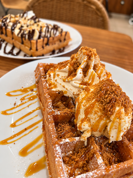 Belgian waffles topped with whipped cream, caramel sauce and cookie crumbs, with another chocolate-covered waffle behind
