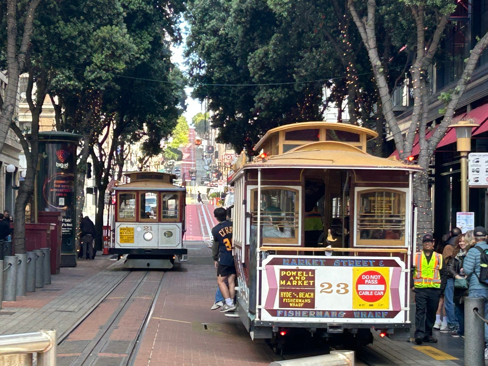 Two historic San Francisco cable cars on Powell Street with tracks, trees, and a steep city hill in the background
